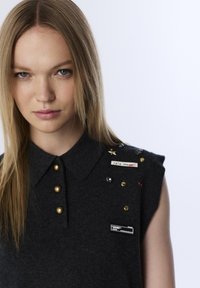 Graues ärmelloses Hemd mit Kragen, verziert mit mehreren dekorativen Anstecknadeln und Knöpfen, aus strukturiertem Stoff gefertigt.