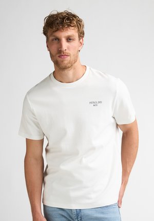 Homme blond avec barbe portant un T-shirt blanc à manches courtes avec le texte "PETROL IND & CO" et un jean bleu clair, debout devant un fond clair uni.