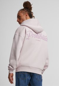 Felpa rosa chiaro con cappuccio, con una grande scritta "Proibito" stampata sul retro. Vestibilità classica e polsini a coste. Jeans in denim visibili.