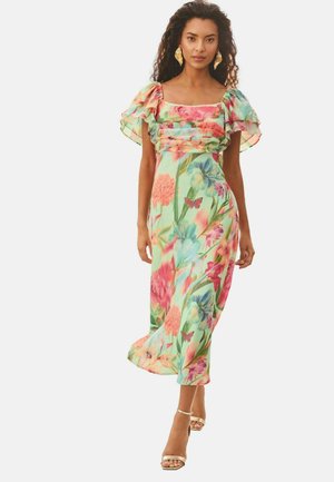 Mujer con un vestido midi floral de colores vivos, mangas abullonadas con volantes y pendientes dorados, caminando con zapatos dorados de punta abierta sobre un fondo liso.