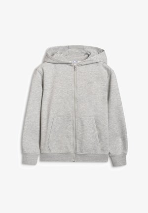 Jeune fille portant une casquette noire, un crop top rose, un sweat à capuche gris clair zippé et un pantalon de jogging assorti, des baskets noires, debout les mains dans les poches.
