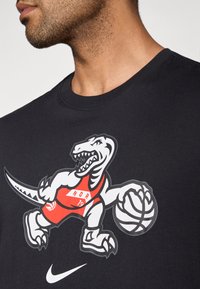 Μαύρο μπλουζάκι με καρτούν δεινόσαυρο σε κόκκινη φανέλα που κρατάει ένα μπάσκετ. Το σχέδιο περιλαμβάνει έντονες γραμμές και ζωντανά χρώματα, με το λογότυπο της Nike.