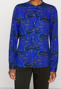 Blue patterned blouse with long sleeves and a mandarin collar, featuring abstract designs in varying shades of blue and dark accents.