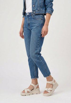 Vrouw met opgerolde blauwe spijkerbroek, spijkerjasje en beige platformsandalen met enkelbandjes, staand tegen een witte achtergrond.