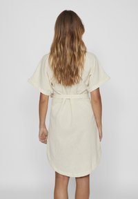 Svijetlo bež lanena haljina s kratkim rukavima i pojasom na vezanje, s zaobljenim rubom i jednostavnim, čistim linijama. Mekana tekstura.