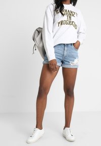 Osoba v bílém svetru s nápisem "Carhartt Work in Progress", roztržených džínových šortkách, bílých teniskách a nese světle šedý batoh.