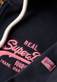 Felpa navy con logo ricamato rosa "Superdry" e coulisse bianca. Presenta un'etichetta di stoffa rosa con design di foglia vicino al collo.