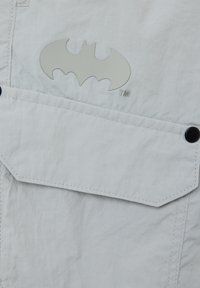 Tela blanca con un acabado texturizado que presenta un gran logo gris de Batman en un bolsillo con cierre a presión. Se aprecian detalles de costura.