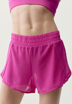 Middenstuk van een vrouw met een strakke buik en roze sportshorts met elastische tailleband tegen een neutrale achtergrond.
