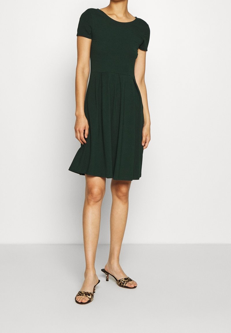 Abito verde scuro fino al ginocchio con maniche corte, corpino aderente e gonna svasata. Indossato con sandali con tacco in stampa leopardata.