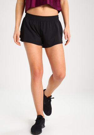 Kvinde iført sorte træningsshorts, sorte sneakers og en kort mørkelilla top, stående med det ene ben let fremad.