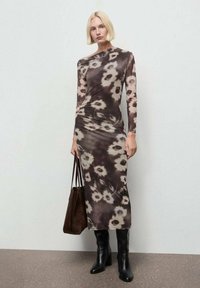 Rochie cu mânecă lungă, ajustată, în nuanțe de brown și crem cu un model floral tie-dye, realizată dintr-un material neted. Este asortată cu cizme negre până la genunchi și o geantă maro.