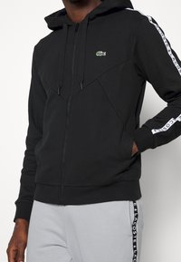 Svart zip-up hoodie med huva, med en krokodil-logotyp, geometriska sömmar och vit Lacoste-logotyp längs ärmarna.