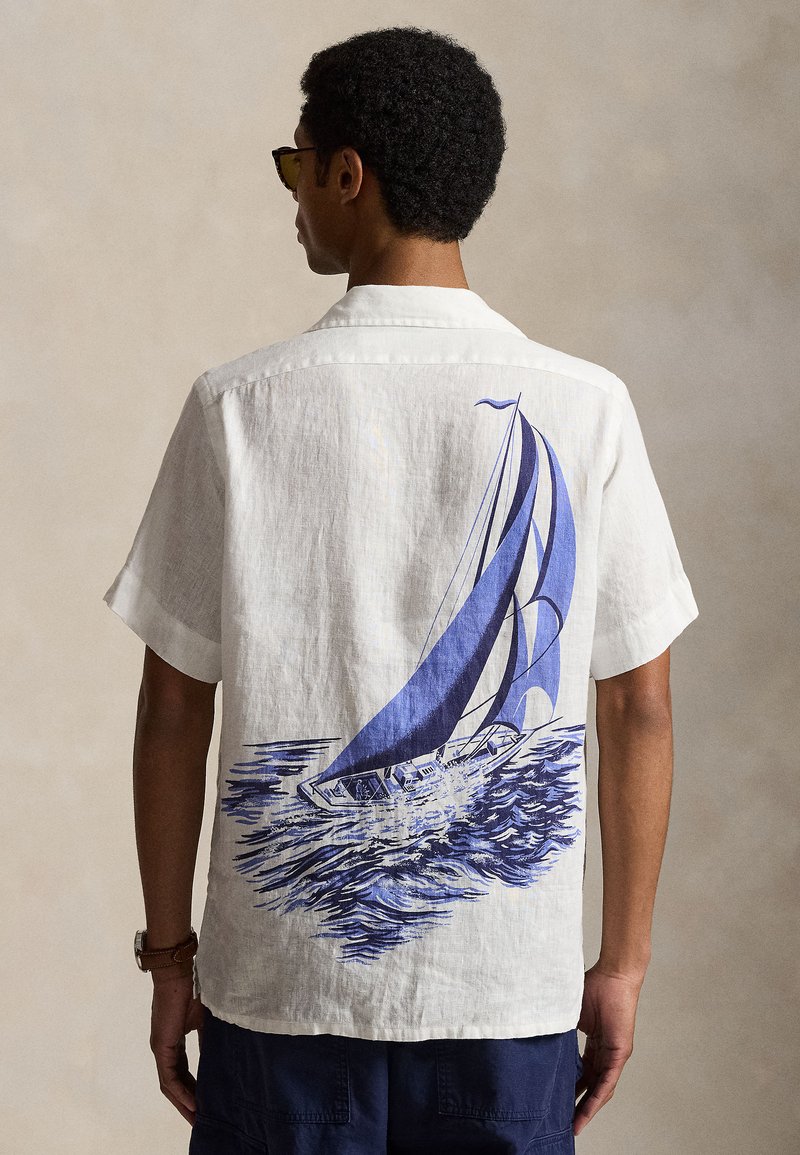 Homme portant une chemise blanche à manches courtes avec un grand motif de voilier bleu dans le dos, associé à un short foncé et des lunettes de soleil.