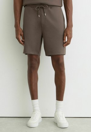 Uomo con pantaloncini marroni con coulisse, calzini bianchi alla caviglia e scarpe da ginnastica bianche, in piedi di fronte a una parete bianca liscia.
