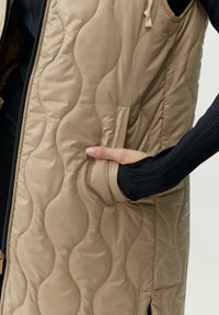 Beige vadderad väst med en slät yta, med en sidoficka och dragkedjestängning. Den inre ärmen visar en åtsittande svart ärm.