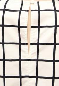 Tissu blanc avec motif à carreaux noirs comprenant une ouverture fendue verticale sécurisée par un seul bouton blanc en haut.