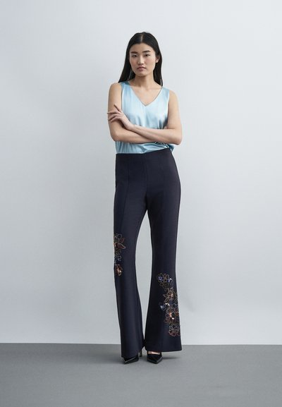 Γυναίκα στέκεται με σταυρωμένα τα χέρια, φοράει έναν ανοιχτό μπλε αμάνικο κορυφή και μαύρο παντελόνι με φαρδιά μπατζάκια με κεντητά λουλουδένια σχέδια.