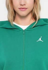 Sudadera con cremallera verde de tejido texturizado, que presenta un logo de Jordan en blanco en el lado izquierdo del pecho y una capucha con un diseño deportivo.