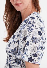 Blumendruckkleid mit blauen und marineblauen Designs auf einem weißen, strukturierten Stoff, mit kurzen Ärmeln und einem gerafften Taillendetail.