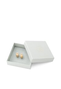Boucles d'oreilles dorées au design géométrique lisse, présentées dans une boîte à bijoux texturée gris clair avec un logo en relief.