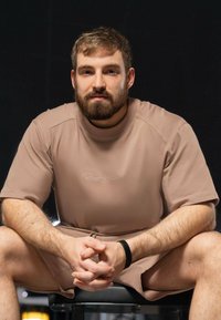 Chemise à manches courtes beige avec une texture subtile et un logo sur la poitrine, associée à un short assorti. Teint de peau lisse, assis avec les mains jointes.