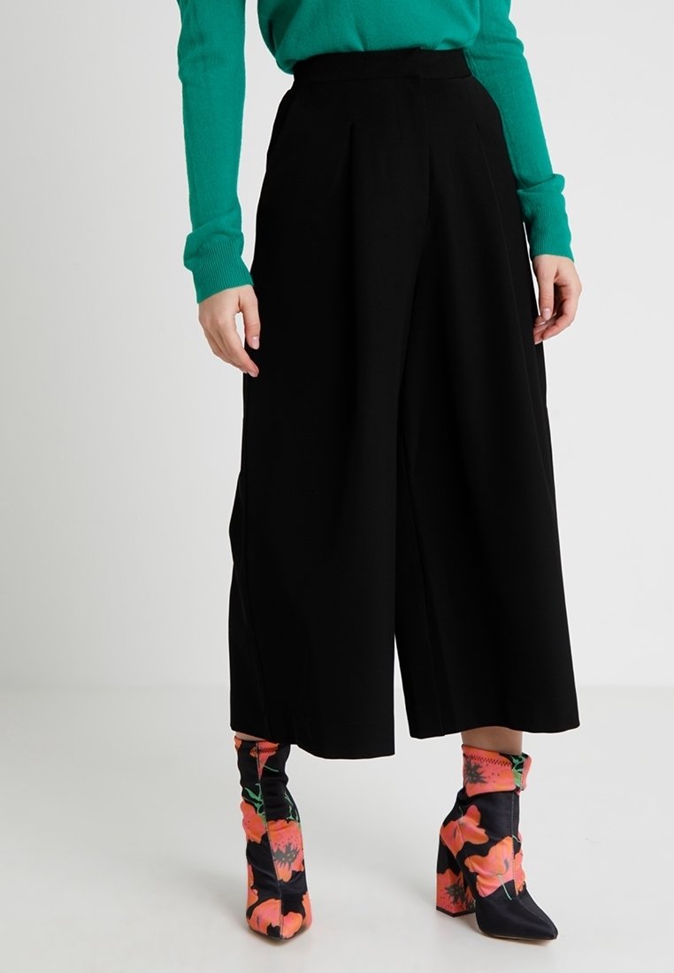 Pantalon noir à jambes larges avec des plis, associé à des bottines aux motifs floraux vibrants avec un talon épais. Pull vert visible.