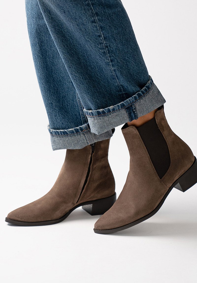 Hnědé semišové kotníkové boty se špičatou špičkou, elastickými bočními panely a nízkým blokovým podpatkem, spárované s krátkými džínami.
