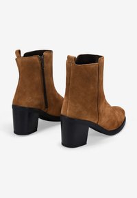 Botines de ante marrón con punta redondeada, tacón alto en bloque y cremallera lateral. Textura suave, diseño minimalista y una lengüeta en la parte trasera.