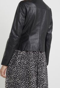 Černá kožená zkrácená bunda s hladkým povrchem, se špičatým límcem a viditelnými švy, vrstvená přes vzorované šaty.