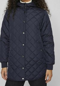 Granatowa, pikowana długa kurtka z kapturem, zatrzaskami oraz dwiema bocznymi kieszeniami; posiada wzór w romby i ściągacze na rękawach.