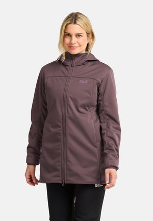 Donna che indossa una lunga giacca viola con zip e collo alto, con le mani rilassate lungo i fianchi, su sfondo bianco uniforme.
