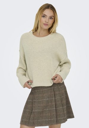 Mujer con pelo rubio que lleva un suéter de punto crema y una falda plisada marrón con estampado, posando con las manos cerca del dobladillo del suéter.