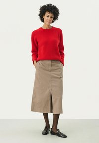 Pull en maille côtelée rouge à manches longues, associé à une jupe beige à longueur genoux avec une fente avant. Des chaussures plates noires complètent la tenue.