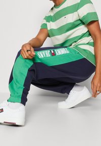 Randig grön och vit t-shirt i kombination med marinblå och gröna joggers med en logotyp. Vita sneakers fulländar den avslappnade outfiten.