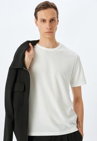 Vit kortärmad texturerad t-shirt med rund halsringning, tillsammans med en svart jacka hängande över axeln, mot en neutral bakgrund.