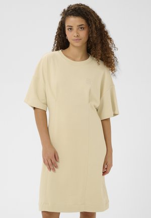 Ung kvinde med krøllet hår iført en løs beige t-shirtkjole, der går til knæene, stående foran en ensfarvet lys grå baggrund.