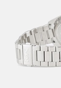Pulsera de reloj de acero inoxidable plateado con un acabado pulido, que presenta eslabones rectangulares y un cierre grabado con "MK" y "MICHAEL KORS."