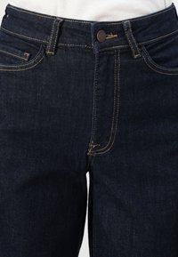 Temno modri jeans z visokim pasom, z zlatimi šivi, kovinskim gumbom za zapiranje in sprednjimi žepi. Gladka tekstura in klasičen kroj.