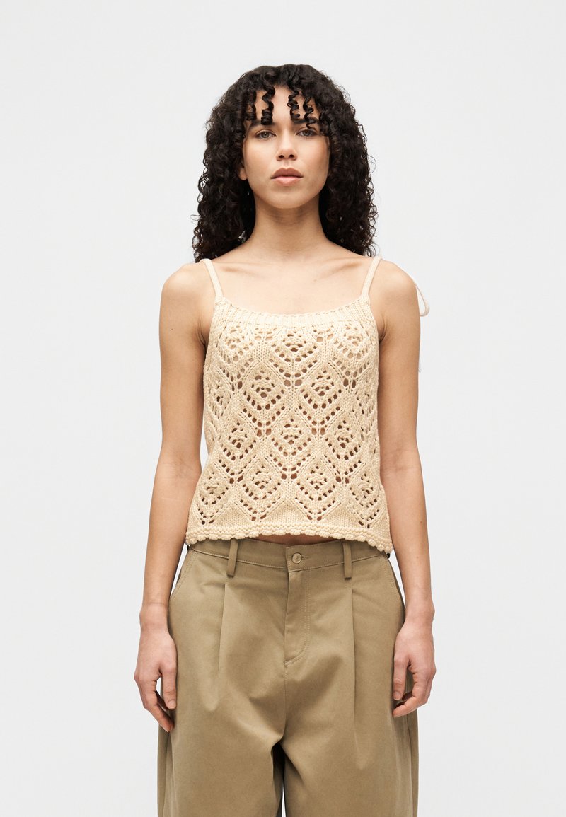 Μπεζ πλεκτό αμάνικο τοπ με διαμάντι μοτίβο και κυματιστό στρίφωμα, φορεμένο πάνω από χαλαρά μπεζ παντελόνια. Διαθέτει λεπτές ρυθμιζόμενες τιράντες.