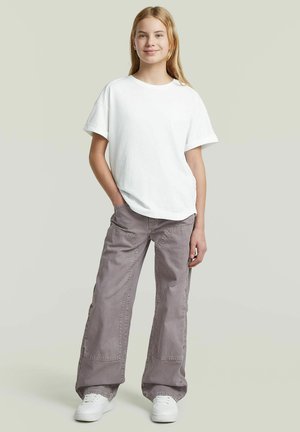 Jeune personne debout, les mains dans les poches, portant un t-shirt blanc, un pantalon gris ample et des baskets blanches sur un fond uni.