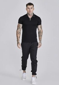 Sort polo t-shirt med lynlåsåbning ved kraven, båret med sort, tilspidsede bukser og hvide sneakers. Outfittet har et figursyet design og glat tekstur.