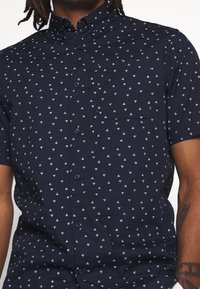 Camicia button-up a maniche corte, di colore blu navy scuro, con motivi a triangolo bianchi, una tasca sul petto e un design con colletto classico.