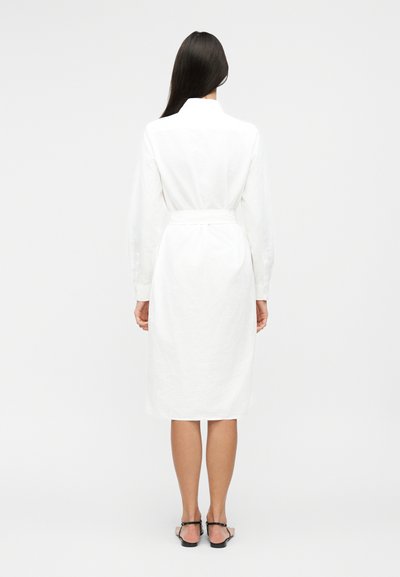 Femeie cu păr lung și întunecat, purtând o rochie midi albă cu mâneci lungi și curea în talie, și sandale negre plate, stând cu spatele la privitor pe un fundal simplu.