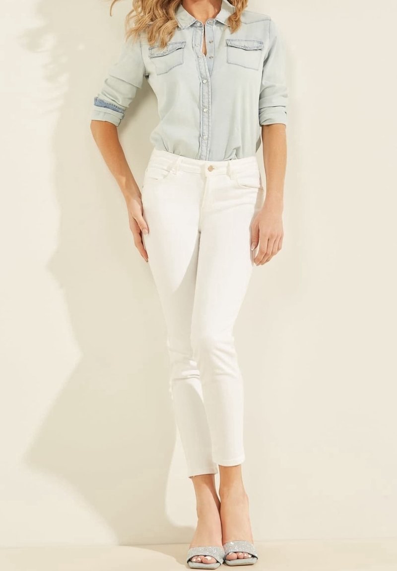 Femme portant une chemise bleu clair boutonnée, un pantalon blanc ajusté et des talons ouverts argentés, debout devant un fond clair.
