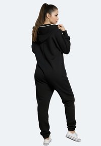 Schwarzer Kapuzen-Overall aus weichem Stoff mit langen Ärmeln und Beinbündchen. Verfügt über einen Reißverschluss und kontrastierende Nähte.