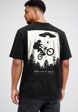 Man wearing black t-shirt with black and white graphic of cyclist jumping and a UFO abducting a person above forest trees.