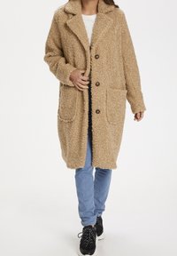 Abrigo marrón claro y esponjoso con cuello con muesca, dos bolsillos delanteros y botones marrones, llevado sobre una camiseta blanca y vaqueros azules con zapatillas negras.