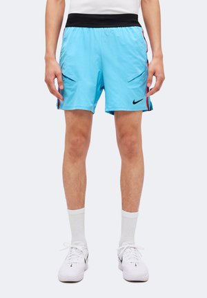 Homme portant un short de sport bleu clair avec une ceinture noire, des chaussettes blanches et des baskets blanches Nike, debout devant un fond uni.