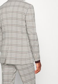 Grijs geruit pak met een gestructureerde stof, met zwarte en rustkleurige accenten. De achterkant toont een moderne pasvorm en details met knoopmanchetten.